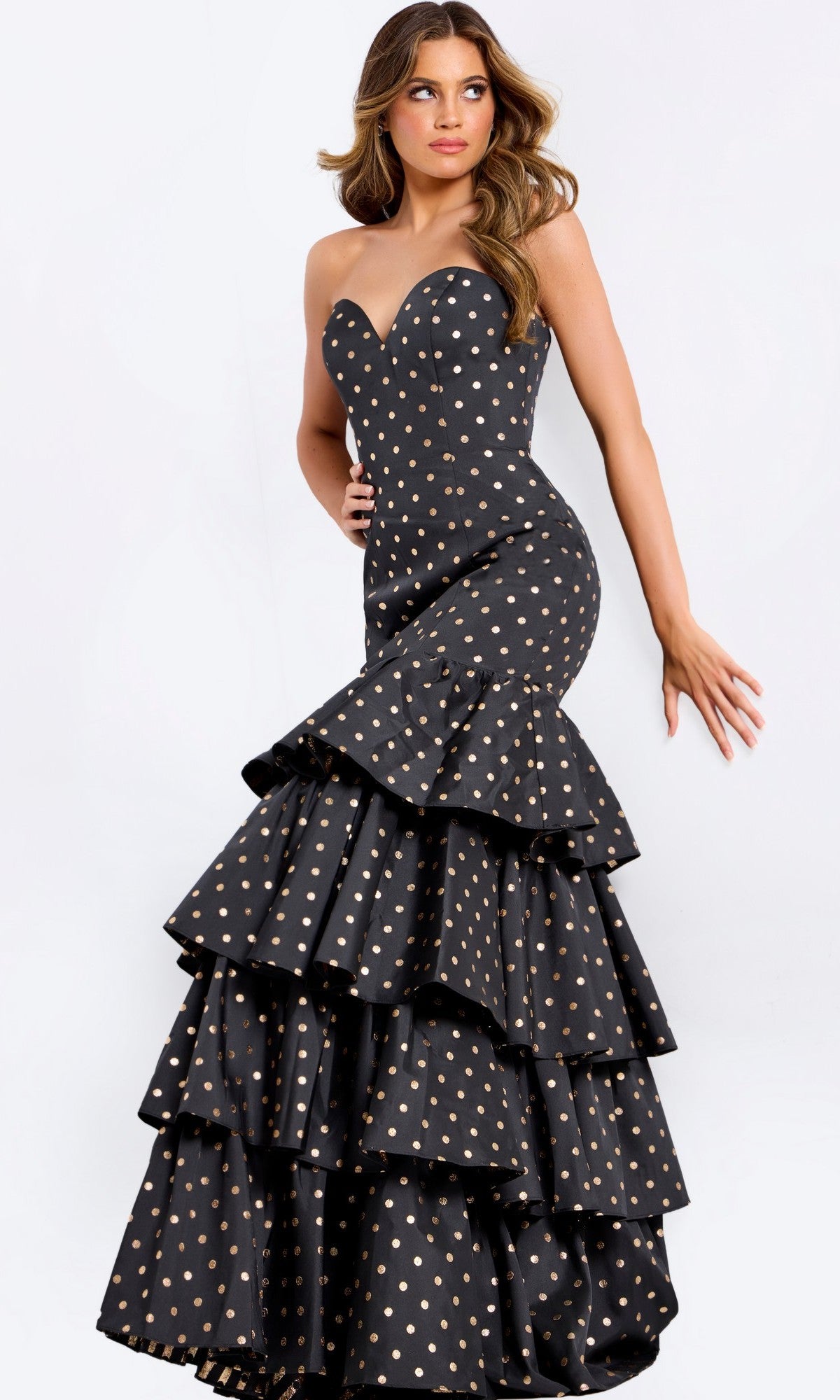 A woman poses against a plain light background in the elegant Jovani 49419 Long Prom Dress, featuring a strapless mermaid silhouette in black/gold with gold polka dots and a tiered ruffled skirt for timeless glamour.